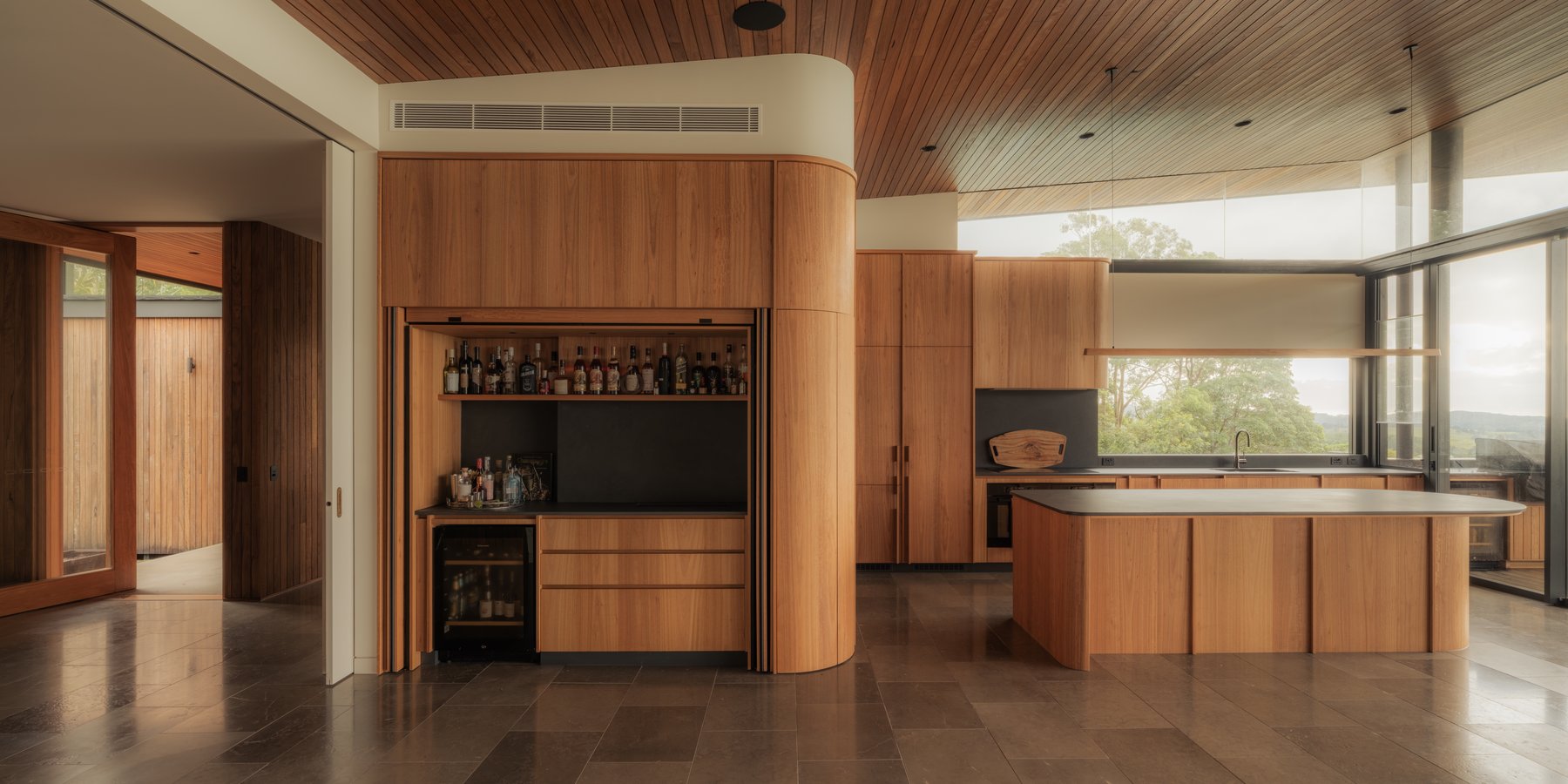 Curved timber bar joinery opening onto a kitchen with stone floors and floor-to-ceiling glazing, Newrybar Residence